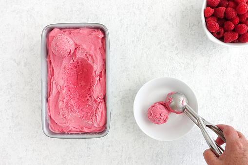 Hand scooping from a container filled with homemade raspberry ice cream and placing a scoop into a bowl.