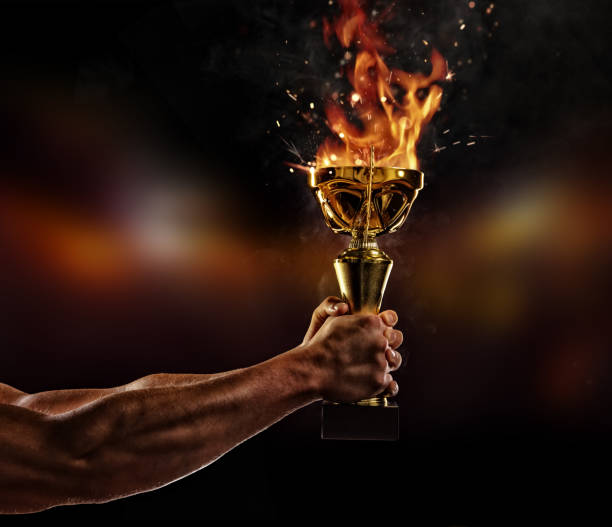 istock Muscular man arm holding burning trophy cup on black background 827881528