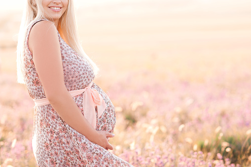Happy pregnant woman wearing stylish dress holding tummy in meadow. Happiness.