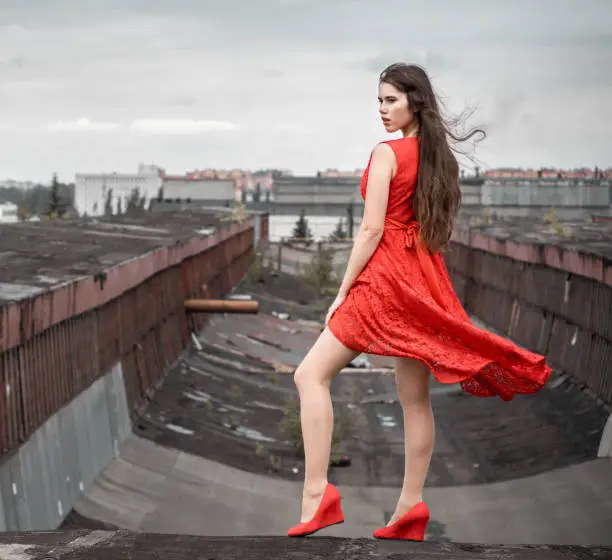 Beautiful young woman with long brown hair in red lace dress stands on the roof a sideways to the camera. She is looking away. The wind develops the hem of woman's dress. Summer shooting at outdoor on old building on a overcast background