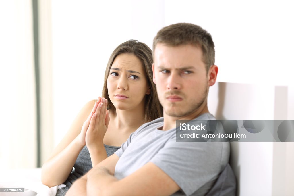 Wife begging and angry husband Wife begging to her angry husband sitting on the bed in a house interior Men Stock Photo