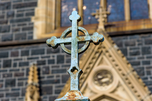 A rusted and weathered iron crucificx in from of a stone wall church with stained glass winndows
