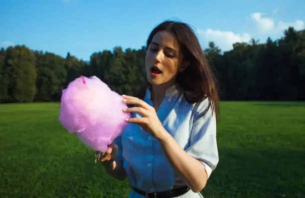 young pretty brunnette woman eating pink cotton candy outdoor on nature park