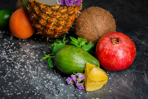 Close-up of tropical fruit assortment on a black stone background. Green avocado, cut carambola, ripe garnet, hard hawaiian coconut and a fresh pineapple. Citruses with mint leaves and purple flowers.