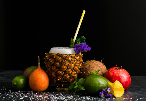 A multi-colored composition of exotic fruits and citruses on a black table background. A big pineapple cup with a striped straw. Refreshing garnet, rustic coconut, green avocado with mint and flowers.