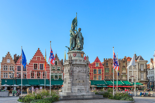Tourists and locals in the Grote Markt -Market Square- of Bruges, a city in the Flemish Region of Belgium. The large town square is lined by notable buildings, and hosts the nineteenth-century monument to Jan Breydel and Peter De Coninck, heroes of the Battle of the Golden Spurs of July 1302.