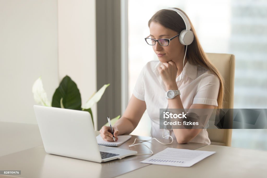 Focused attentive woman wearing headphones using laptop at office desk Focused attentive woman in headphones sits at desk with laptop, looks at screen, makes notes, learns foreign language in internet, online study course, self-education on web, consults client by video Listening Stock Photo