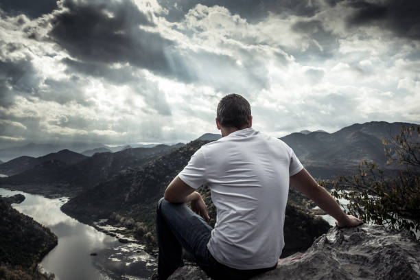 Lonely man looking with hope at horizon on mountain peak with dramatic sunlight during sunset with effect of light at the end of tunnel with moody sky
