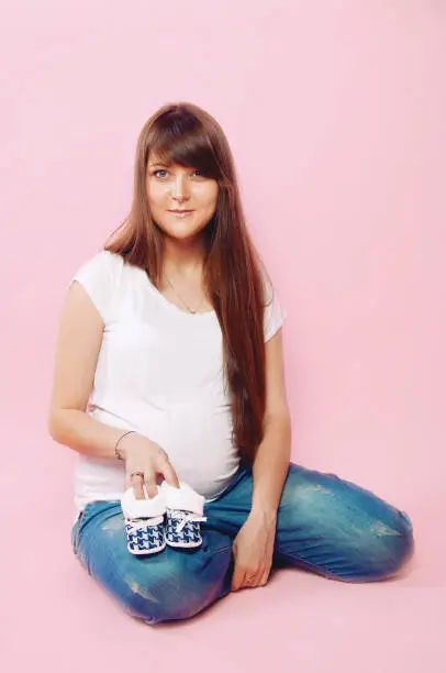 Pregnant woman in jeans and white tee shirt posing on pink background holding baby shoes. Happy mother-to-be. Space for text.