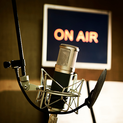 A condenser microphone on a stand in an audio recording booth with an 'On Air' warning illuminated.