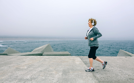 Senior sportswoman with headphones running by sea pier