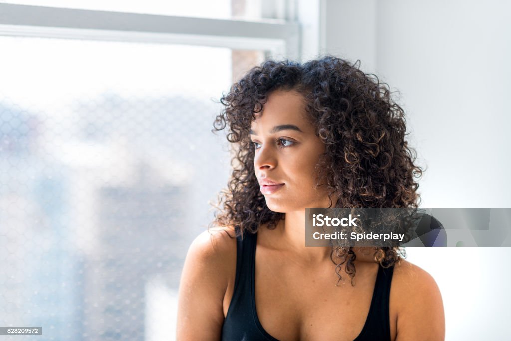 Woman Stares Out of the Window of Her Loft Young adult latin hispanic female sits with head turned profile staring out of the window of her loft or office. One Woman Only Stock Photo