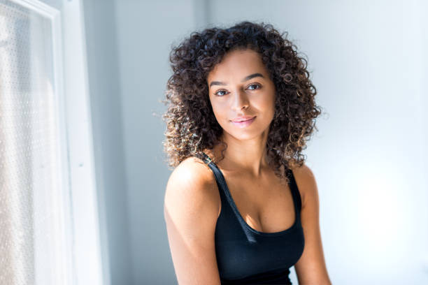 Female Spokesperson Young adult latin hispanic female sits next to window of her loft or office smiling into the camera. cleavage stock pictures, royalty-free photos & images