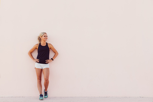 A woman runner stands against a pink wall while stretching before her run. She has her hands on her hips and is smiling because she is confident with her body and mind.