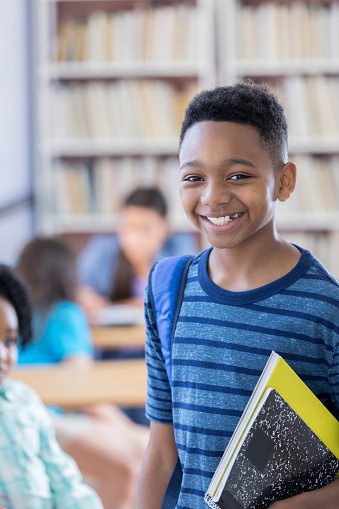 An attractive African American preteen male student smiles for the camera while standing in the library at his school.  He is wearing a backpack on one shoulder and carrying a couple of notebooks under one arm.