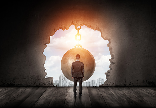 A businessman standing with a turned back and looking into the city through a wall hole made by a giant wrecking ball. Business and opportunity. Assistance and help. Breakthrough.
