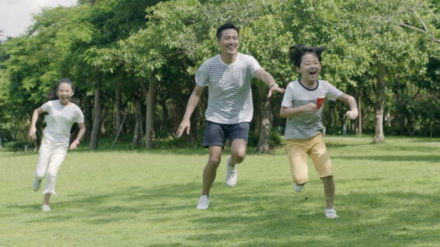 asian father playing chase game with his son & daughter in park in slow motion