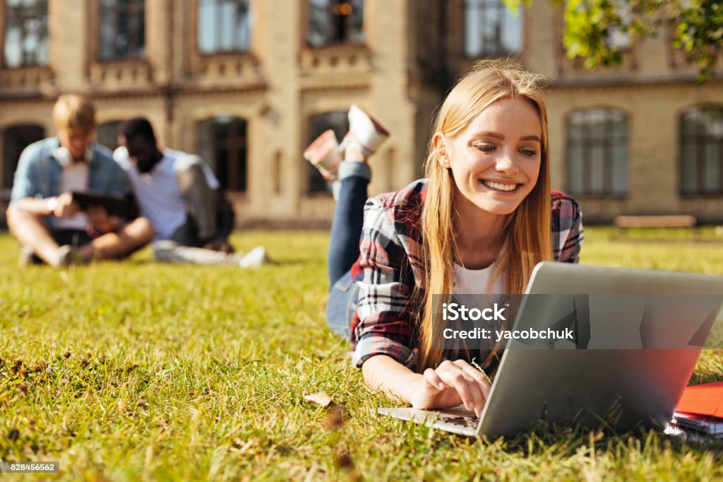 Young optimistic woman checking her updates Internet everywhere. Progressive productive pretty girl lying on the grass and connecting to the free wifi at the campus while reading messages online University Stock Photo