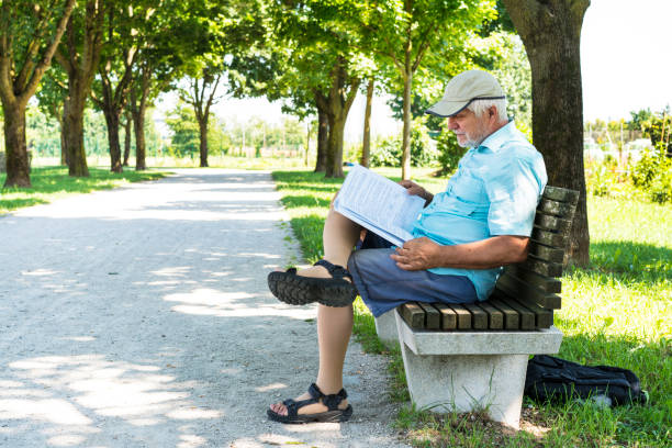 Side view of Senior reading a book while sitting on bench in shadow on The Trail of Remembrance and Comradeship in Ljubljana, Slovenia
