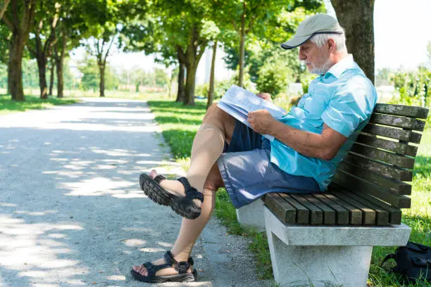 Photo of Side view of Senior reading a book while sitting on bench in shadow on The Trail of Remembrance and Comradeship in Ljubljana, Slovenia