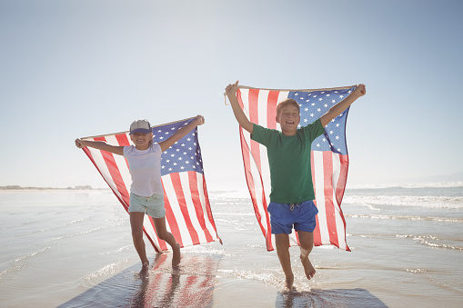 Siblings holding American flags while running on shore at beach during sunny day