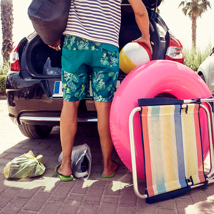 closeup of a young caucasian man putting in or taking out beach stuff from the trunk of his car, such as a beach chair, a pink swim ring or a inflatable beach ball