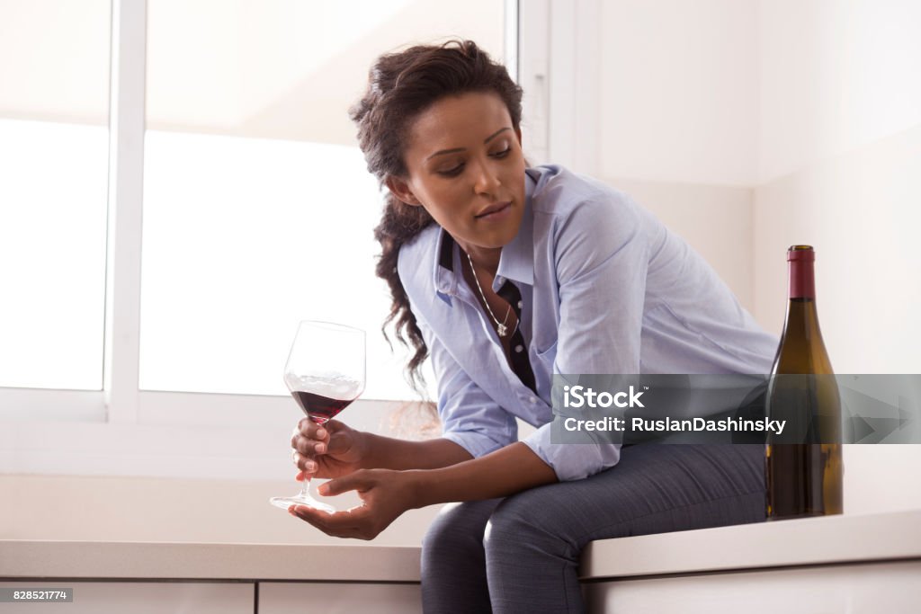 Woman relaxing after work by drinking wine in the kitchen. Pensive business woman sitting on the marble kitchen counter, holding a glass of wine and looking at wine bottle. Woman drinking alcohol, trying to relieve workplace stress. Alcohol Abuse Stock Photo