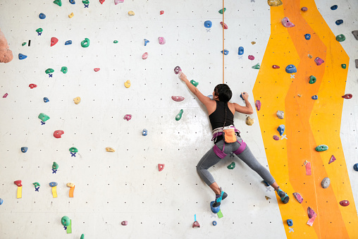 Sportspeople practicing at climbing center. Woman doing rock climbing on artificial climbing wall with instructor standing below holding harness rope.