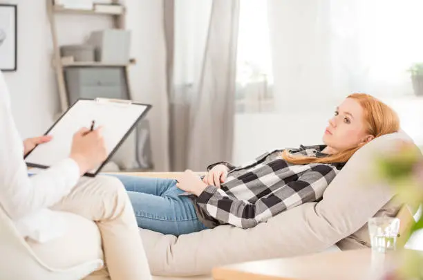 Depressed woman lying on sofa during psychotherapy session, psychologist making notes in medical card