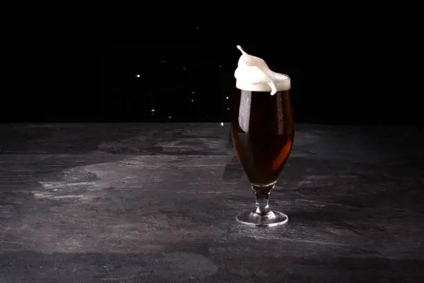 Photo of A big glass of beer full with brown ale and with white foam blowed away on a dark stone background.