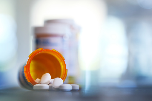 Pills pour out of a prescription medication bottle onto a kitchen counter.  Several other pill bottles stand out of focus in the background.