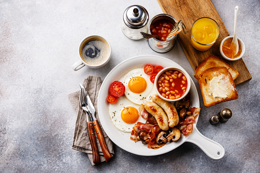 English breakfast in white pan with fried eggs, sausages, bacon, beans, toasts, orange fresh and coffee on white background copy space