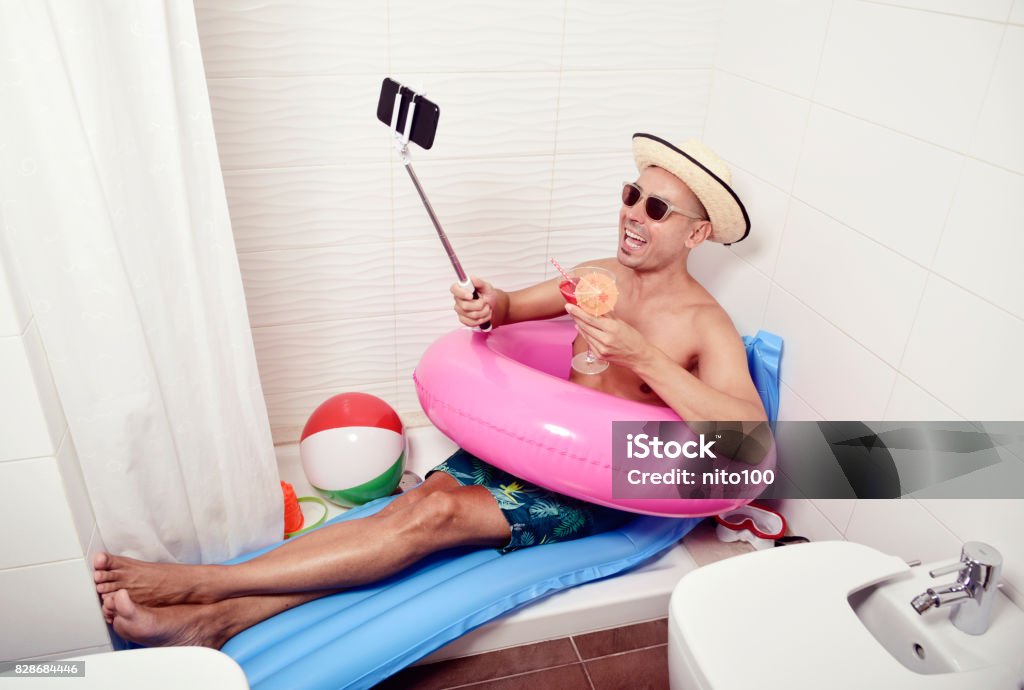 man with swim ring taking selfie in the bathroom a young caucasian man wearing sunglasses, a straw hat and a pink swim ring in a blue air mattress placed in the shower of a bathroom takes a selfie with his smartphone while is drinking a red cocktail Humor Stock Photo