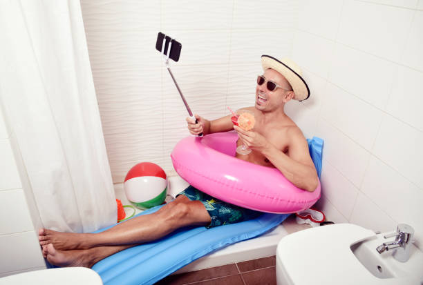 man with swim ring taking selfie in the bathroom a young caucasian man wearing sunglasses, a straw hat and a pink swim ring in a blue air mattress placed in the shower of a bathroom takes a selfie with his smartphone while is drinking a red cocktail staycation photos stock pictures, royalty-free photos & images