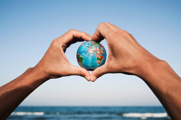 young man with a world globe in his hands closeup of a young caucasian man with a world globe in his hands forming a heart against the blue sky, in front of the sea earth in hands stock pictures, royalty-free photos & images