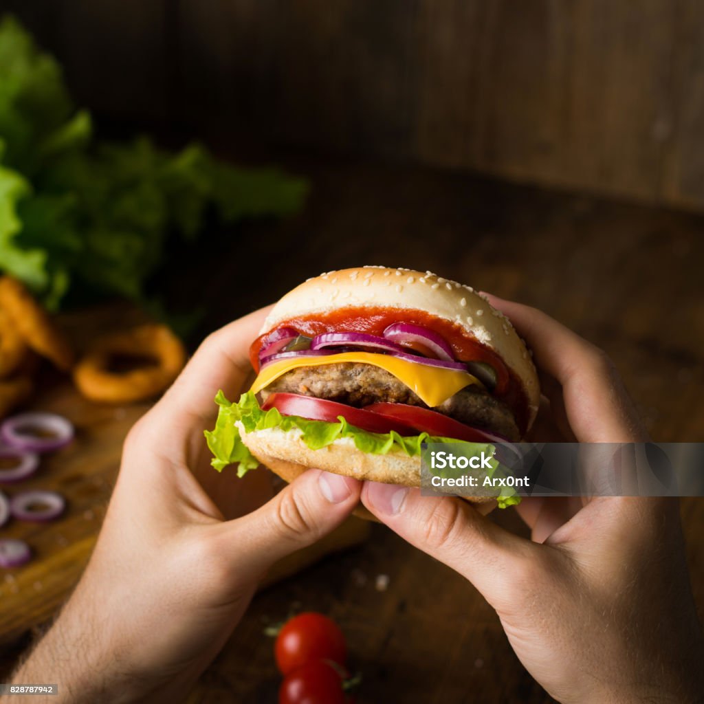 Cheeseburger in hands. Eating cheeseburger Cheeseburger. Man hands holding burger with cheese, red onion, tomatoes, lettuce green salad and pickles. Square crop. Closeup view, selective focus Ketchup Stock Photo