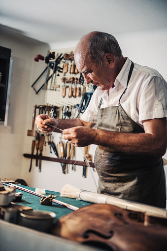 Portrait of senior entrepreneur working in his instrument repair shop, taking care of violins and other music instruments. Experienced senior is using his skills to repair instruments