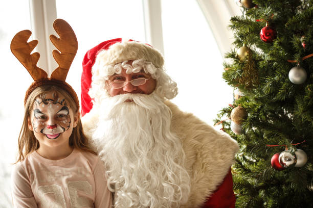 Toy portrait of Santa Claus in glasses and a red hat hanging on the Christmas tree branch, white blinds background