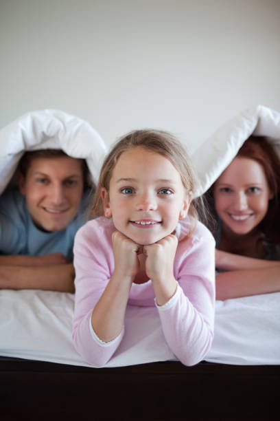 istock Smiling girl under bed cover with her parents 829218036