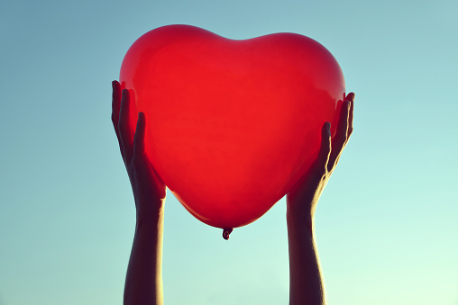 Hands holding red balloon in the shape of heart at sunset.