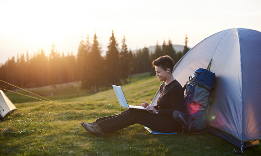 Cheerful young female camping outdoors sitting near her tent using laptop