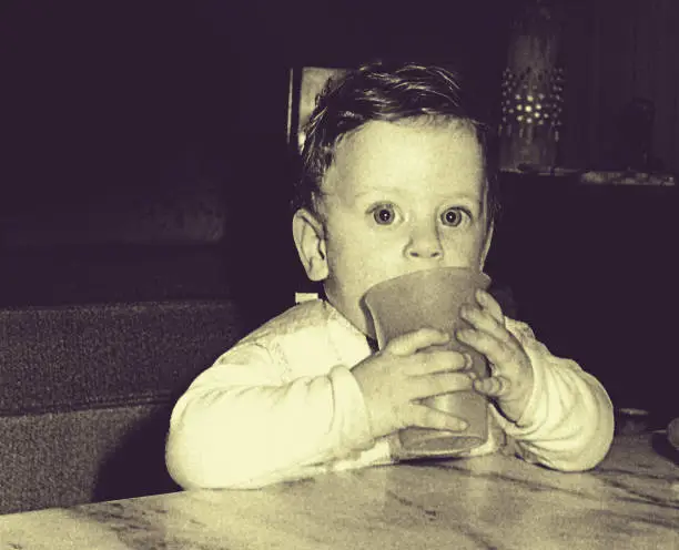 Vintage photo of a baby from the sixties drinking water from a glass whie looking at camera