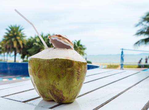 Coconut water on white wooden table, against blurred beach background in sunny day.