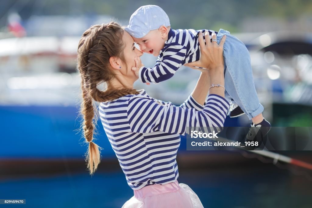 Mother and her little son walking together along the waterfront A young beautiful woman, a brunette with long hair braided in two pigtails, is dressed in a pink transparent skirt and a sea vest, wears small earrings in her ears, plays outdoors in the summer on the dock with her little son dressed in a sea vest Adult Stock Photo