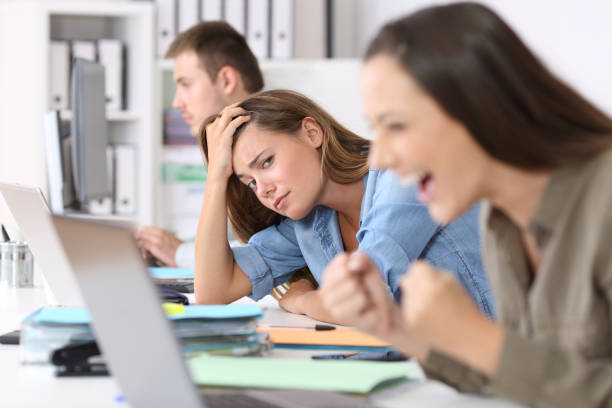 Worried worker beside successful one Worried worker beside a successful one who is excited reading good news on line at office envy stock pictures, royalty-free photos & images