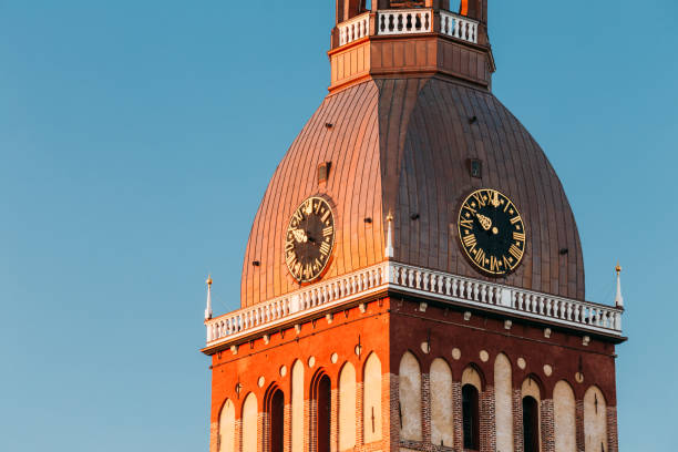 Close Up Of Clock On Tower Of Riga Dome Cathedral In Riga, Latvia. Sunny Blue Sky Background. Tower In Golden Hour At Sunset Or Sunrise TimeClose Up Of Clock On Tower Of Riga Dome Cathedral In Riga, Latvia. Sunny Blue Sky Background. Bellfry Bell Tower In Golden Hour At Sunset Or Sunrise Time