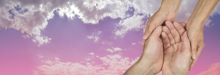 Wide banner with a woman's hands holding a man's cupped hands in a needy gesture on a beautiful pink and blue sky background  with copy space on the left