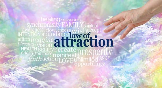 Female hand reaching towards a LAW OF ATTRACTION word cloud  on a multi coloured wispy fantasy style woodland trees and fern  background