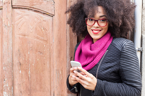 Young african woman with red eyeglasses holding smart phone and looking at camera. Wooden door on background.