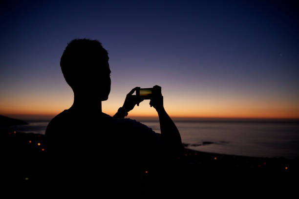 istock Man taking photos with phone on beach, silhouette at sunset 829632478
